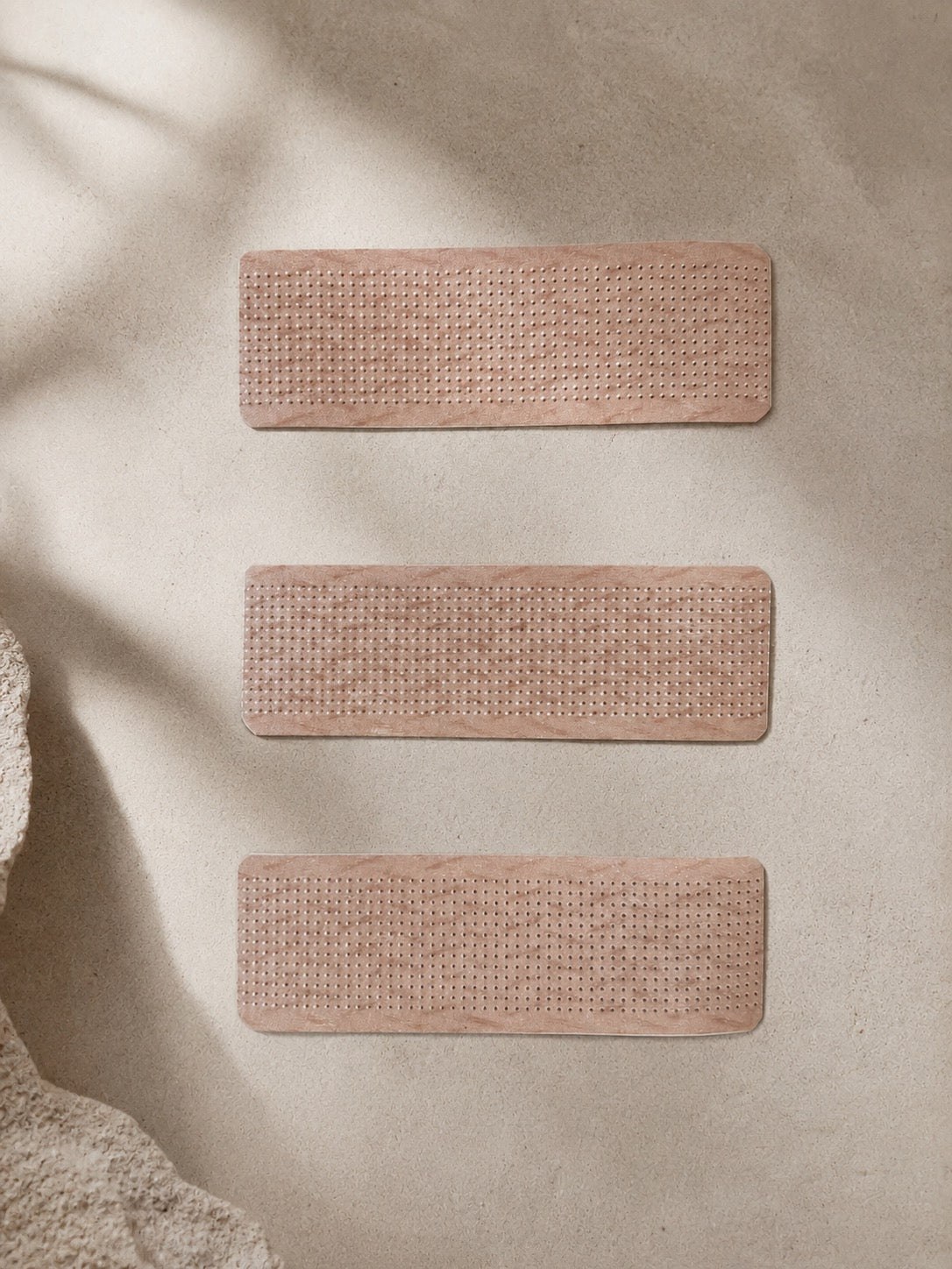 Three rectangular woven placemats in a dusty rose pink color, arranged vertically on a textured beige surface with soft leaf shadows.